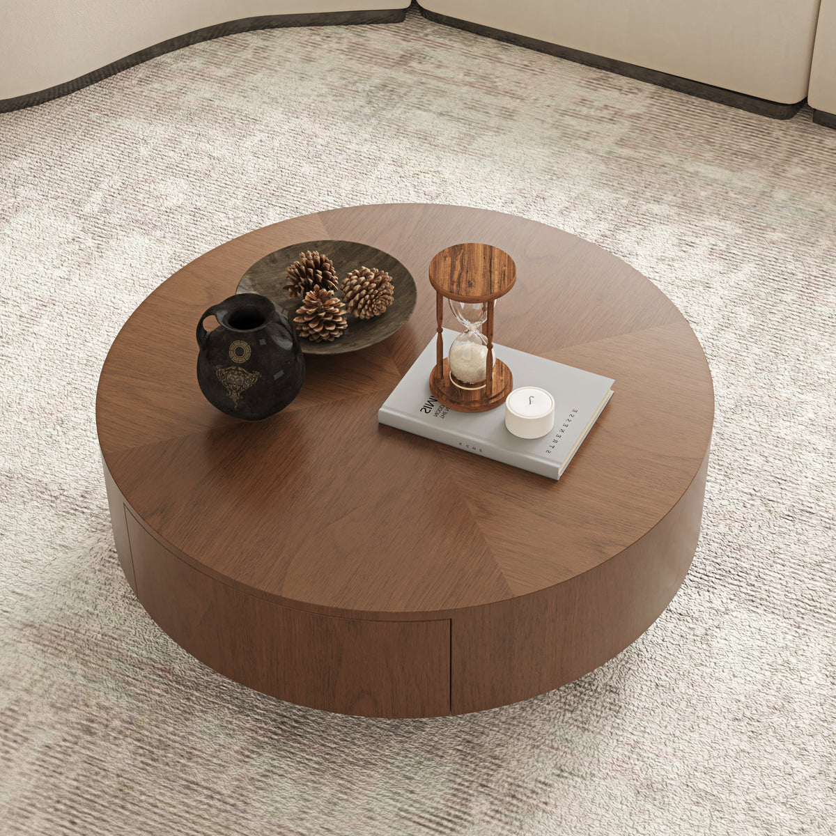 A round wooden coffee table with a warm medium-brown finish, styled with a black ceramic vase, pinecones, a small hourglass, and a candle on a light beige textured rug.