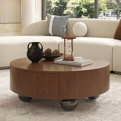 A round wooden coffee table with a warm medium-brown finish and dark metal ball feet, styled in a modern living room setting with a cream sofa, decorative vase, and hourglass.