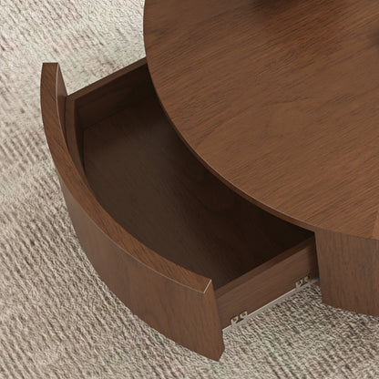Close-up of a round wooden side table with one curved drawer pulled open, showing the wood grain finish and metal pull hardware on a textured light rug.