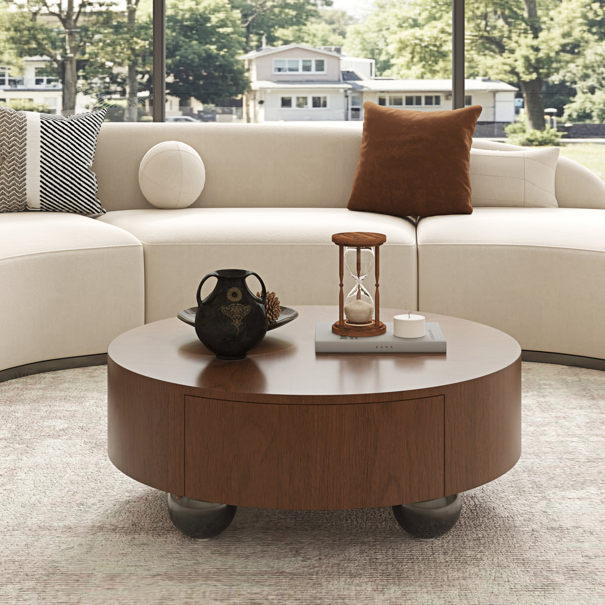 A round wooden coffee table with a dark wood finish and metal legs sits in front of a curved cream sectional sofa. The table is styled with a black decorative vase, a small wooden hourglass, and a candle.