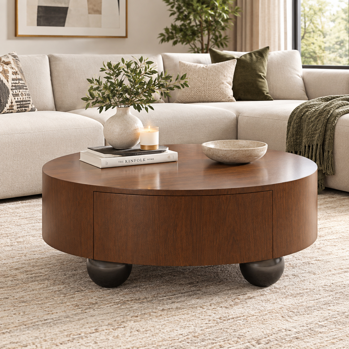 A round wooden coffee table with dark ball feet sits in a modern living room setting with a beige sectional sofa, decorative pillows, a vase with greenery, and a lit candle on top.
