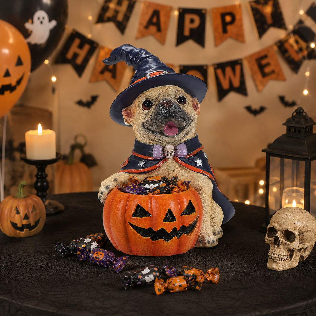 A decorative ceramic pug figurine dressed as a Halloween witch, sitting in a carved jack-o'-lantern candy bowl on a black tablecloth. The scene is decorated with a 'Happy Halloween' banner, candles, a skull, and candy, with the figurine centered in the foreground.