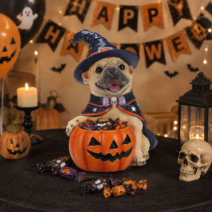 A decorative ceramic pug figurine dressed as a Halloween witch, sitting in a carved jack-o'-lantern candy bowl on a black tablecloth. The scene is decorated with a 'Happy Halloween' banner, candles, a skull, and candy, with the figurine centered in the foreground.